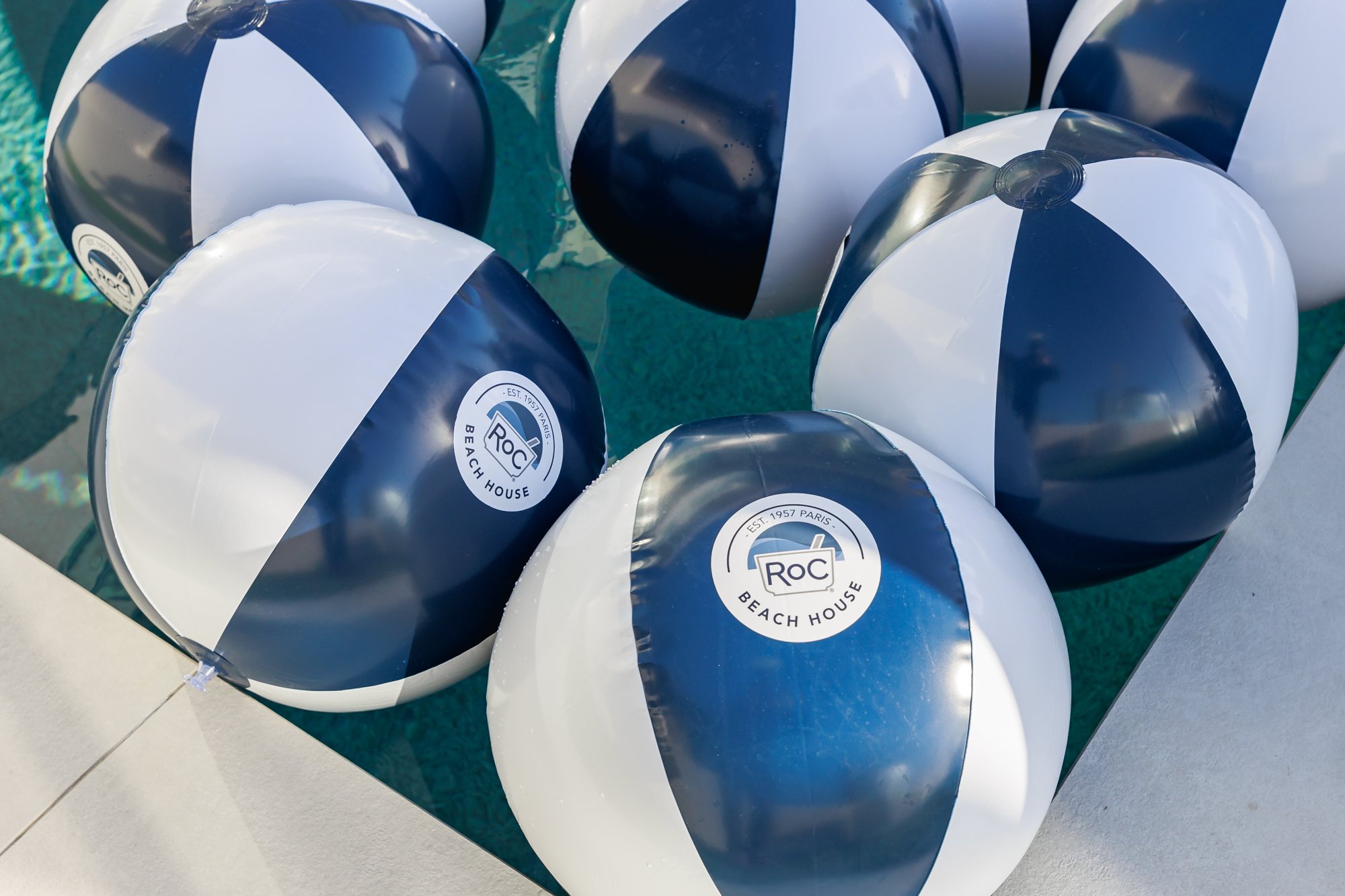 Close-up of multiple beach balls with black, white, and navy blue panels, some displaying a logo for 'RoC Beach House' with a circular design and the text 'Est. 1957 Paris'.