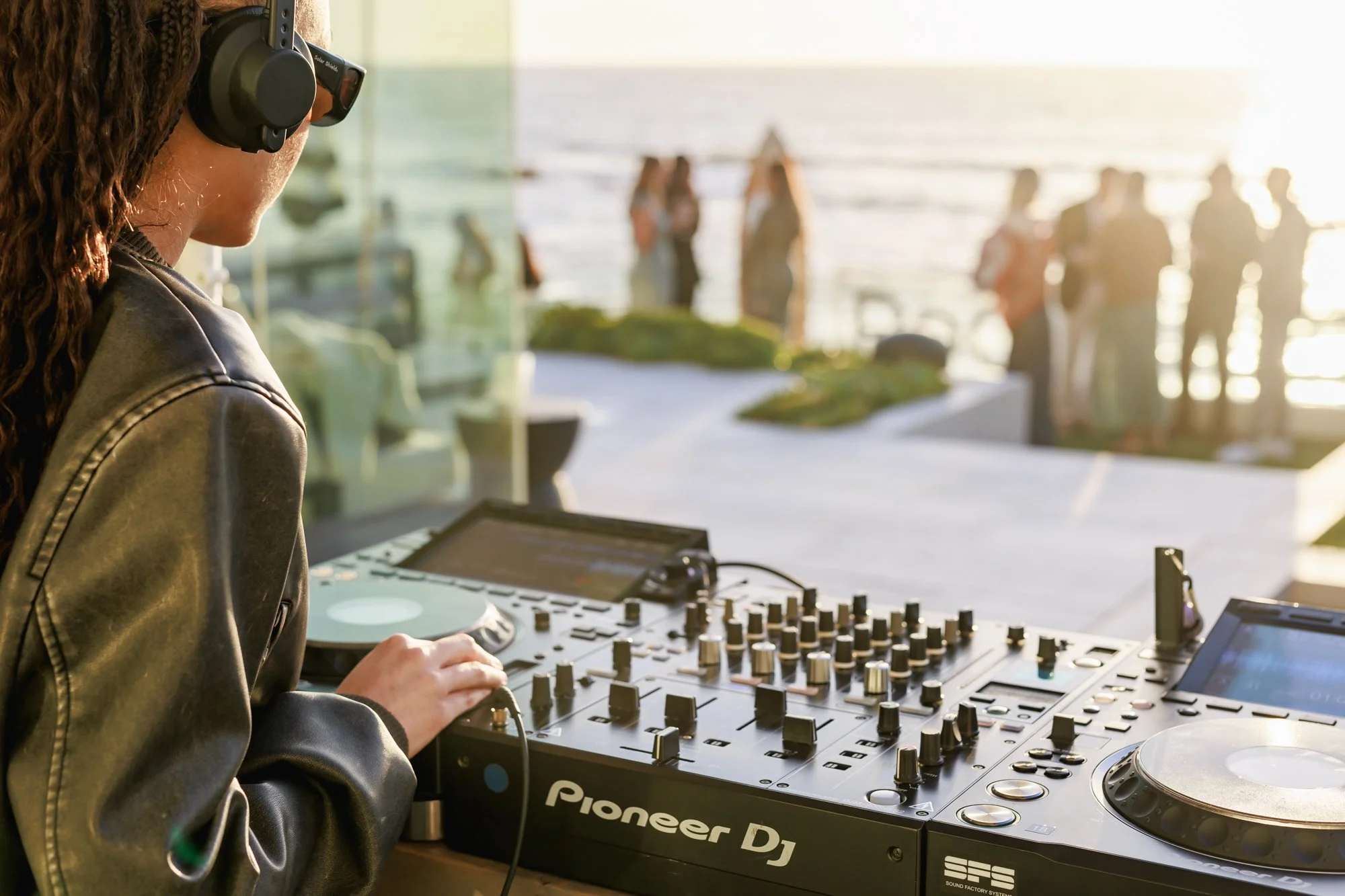 A woman with braided hair wearing headphones, DJing on a Pioneer DJ controller with a beach scene and people in the background.