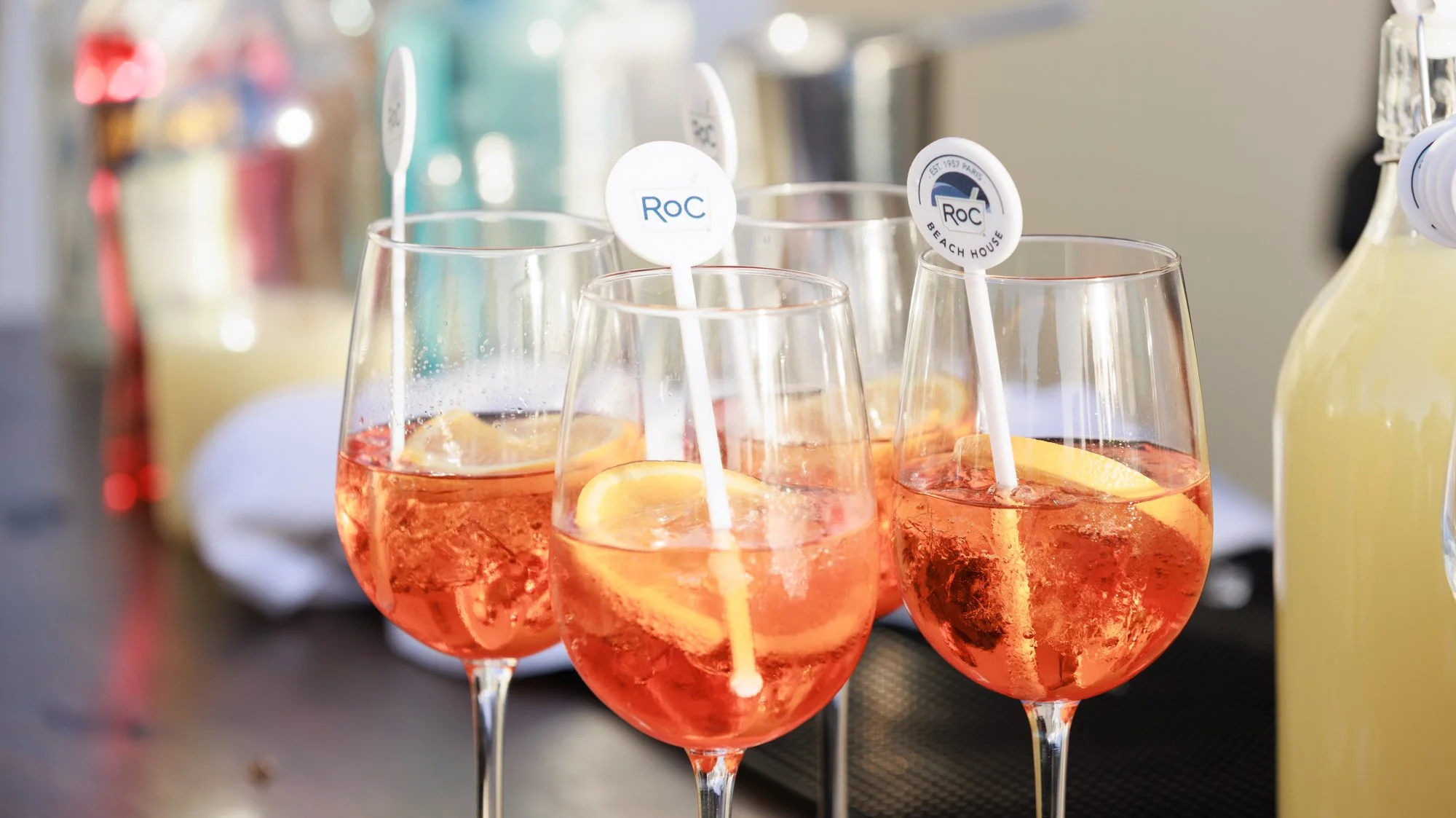 Four glasses of pink-hued cocktail with ice and lemon slices on a black surface with blurred bottles and bar accessories in the background.