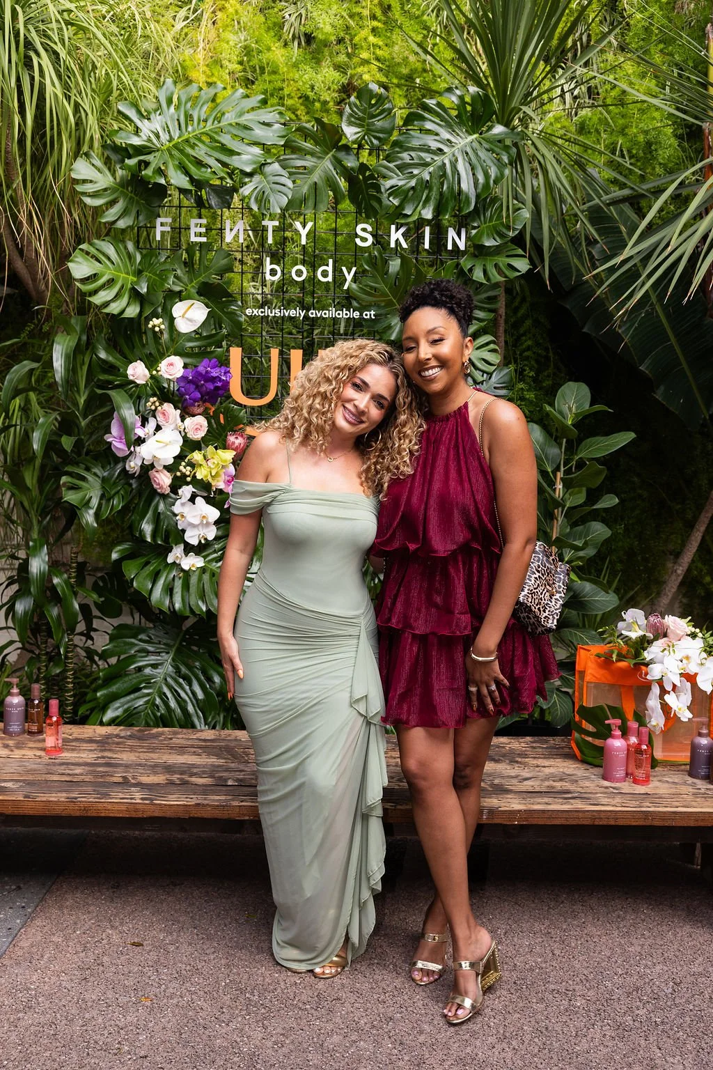 Two women standing close together and smiling in front of a lush green backdrop with flowers and a sign that reads 'Fenty Skin body exclusively available at Ulta.' One woman is wearing a light green off-shoulder dress, and the other is in a burgundy 