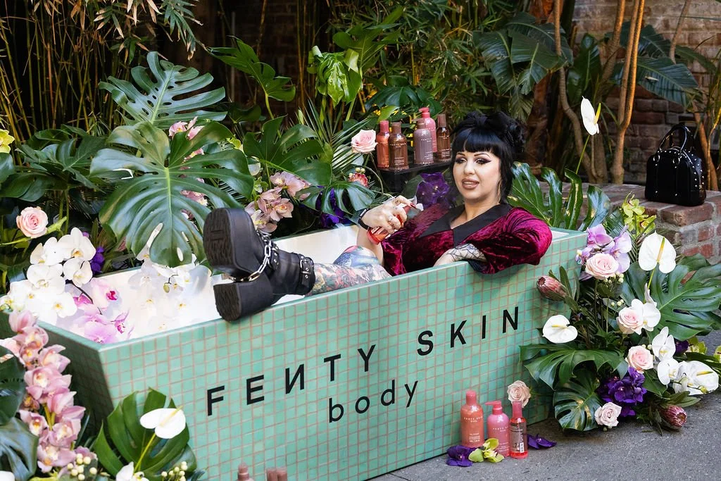 A woman with dark hair and tattoos relaxing in a bathtub filled with flowers and surrounded by green plants, with a sign that says 'Fenty Skin body'.