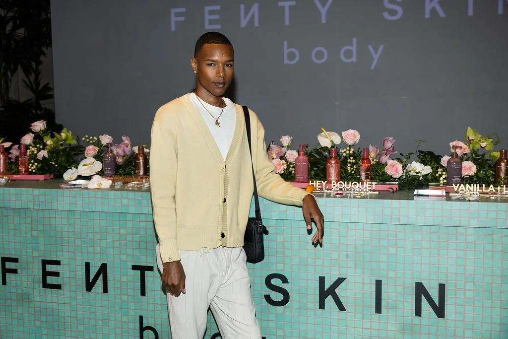 A person stands in front of a Fenty Skin promotional display featuring skincare products and flowers. The person is wearing a cream-colored cardigan, white pants, and a necklace, and is carrying a black shoulder bag.