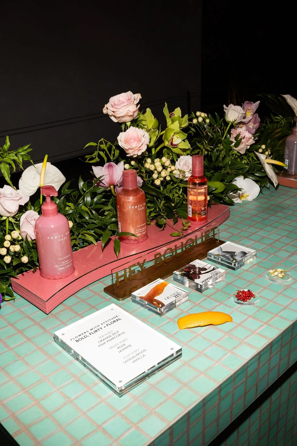 Display of Fenty Beauty skincare products with pink bottles, a floral arrangement with roses and lilies, small plates of berries and petals, and a sign reading 'They Bougie' on a pink-tiled surface.