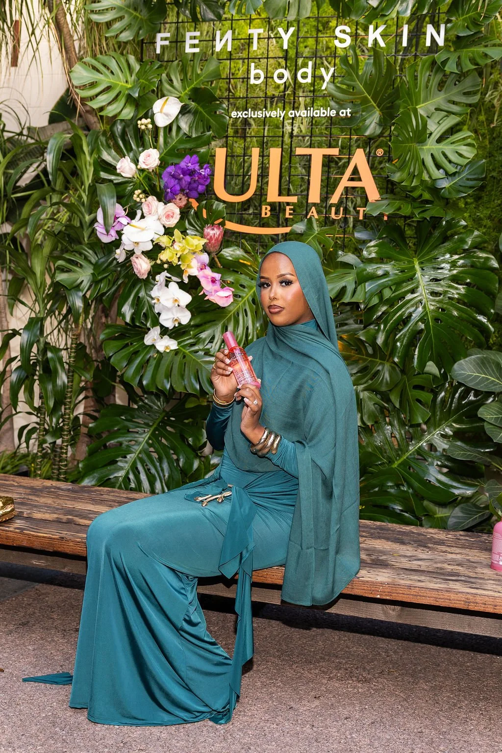 A woman dressed in a teal hijab and matching dress sitting on a wooden bench in front of a lush green backdrop with tropical leaves and flowers. She is holding a pink bottle of skin care product. Behind her, a sign reads 'Fenty Skin body exclusively 