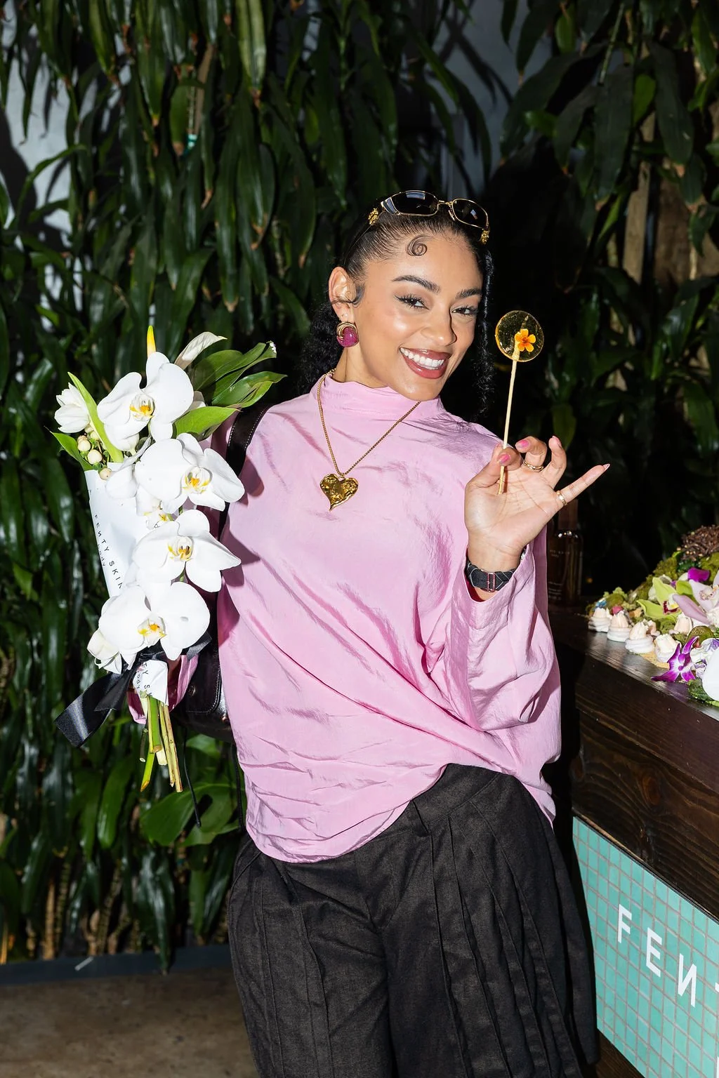 A woman smiling, holding a bouquet of white orchids, standing in front of leafy greenery, wearing a pink top, dark pants, gold jewelry, and sunglasses on her head, with a small dessert and flowers on a table behind her.