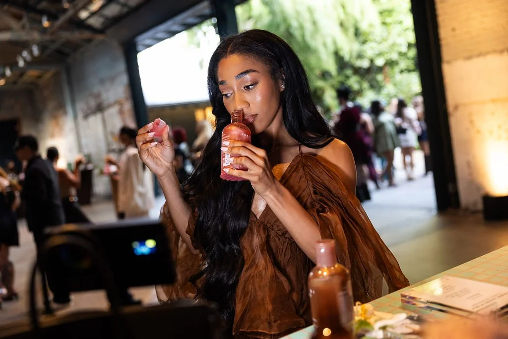 Young woman with long dark hair in brown off-the-shoulder top holding a pink bottle and testing a pink cosmetic product at a social event.