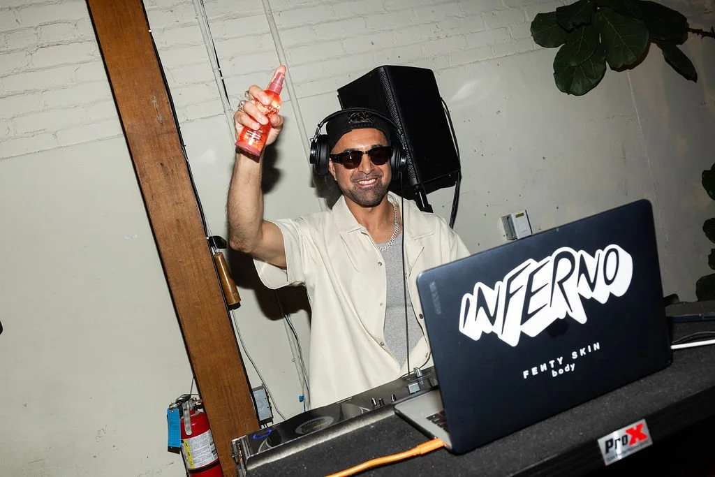 A man DJ wearing sunglasses and headphones, smiling, holding a spray bottle, standing behind DJ equipment with a laptop labeled 'INTERNO' and 'FENTY SKIN body' logo, in an indoor setting.