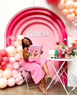Woman sitting on a chair reading a magazine, surrounded by pink and white balloons, with a pink rainbow backdrop and the word 'Caress' in the background, alongside a white table with a typewriter, books, and flowers.