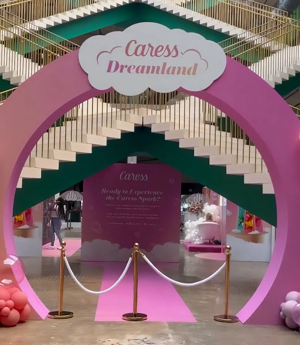 Entrance to a attraction called Cares Dreamland, featuring a pink archway with a cloud-shaped sign reading "Cares Dreamland." Roped-off pathway leading inside, with a pink carpet. Inside, there are displays with pink and white decorations and clouds,