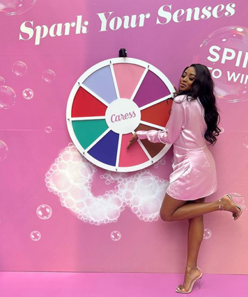A young woman in a pink satin dress stands on one leg next to a colorful spinning wheel labeled 'CARES.' She is smiling and hitting the wheel with her hand against a pink backdrop that says 'Spark Your Senses' and has bubbles and a globe.