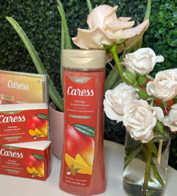 A gift set of Caress Mango & Almond Oil soap in a pink bottle and boxes, displayed next to a bouquet of white roses and pink lilies.