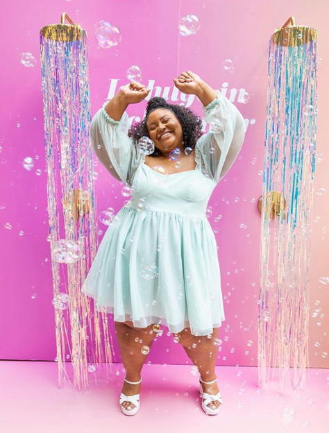 A woman with curly hair and a white dress smiling and raising her arms in front of a pink background with the word 'Inspiring' and decorative shower curtain-like streamers.