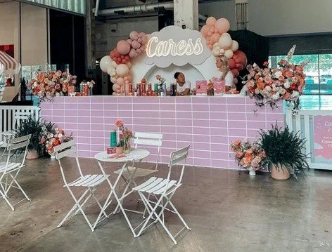 Decorated party table with pink balloons, flowers, and a sign that says 'Caress' in a large indoor space.