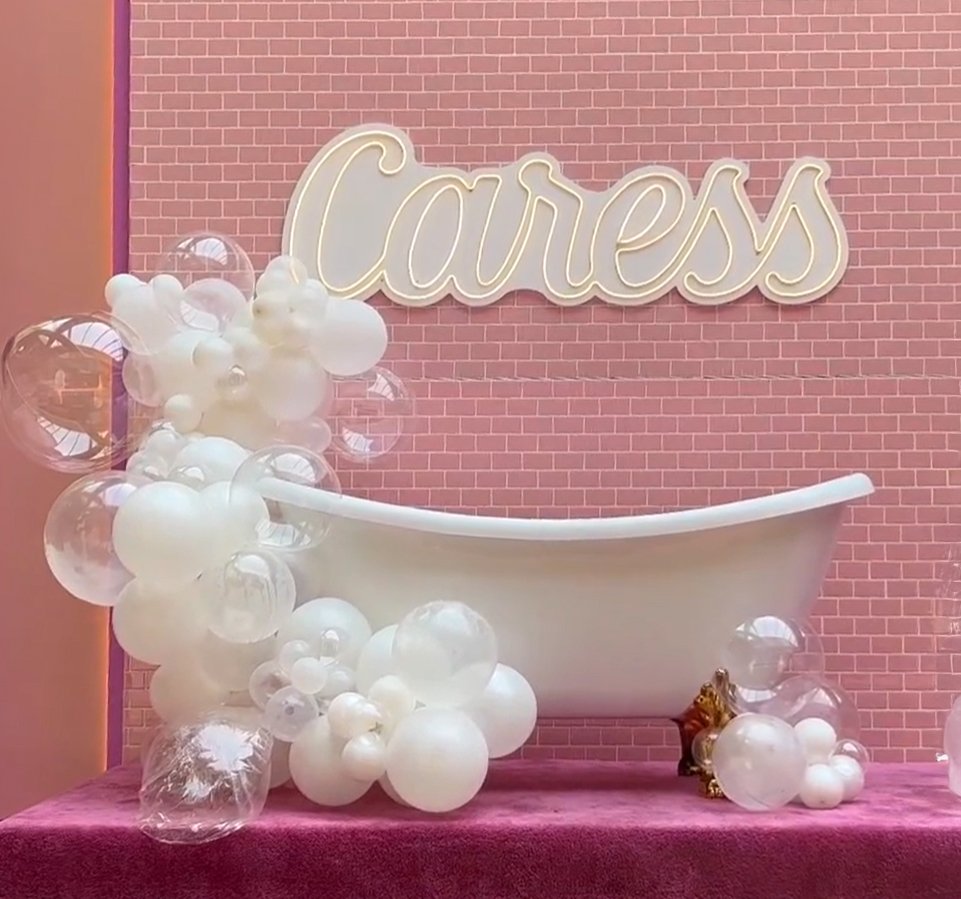 Decorative setup with a white bathtub surrounded by white and clear balloons, pink brick wall background, illuminated 'Caress' sign, and a small golden tiger statue.