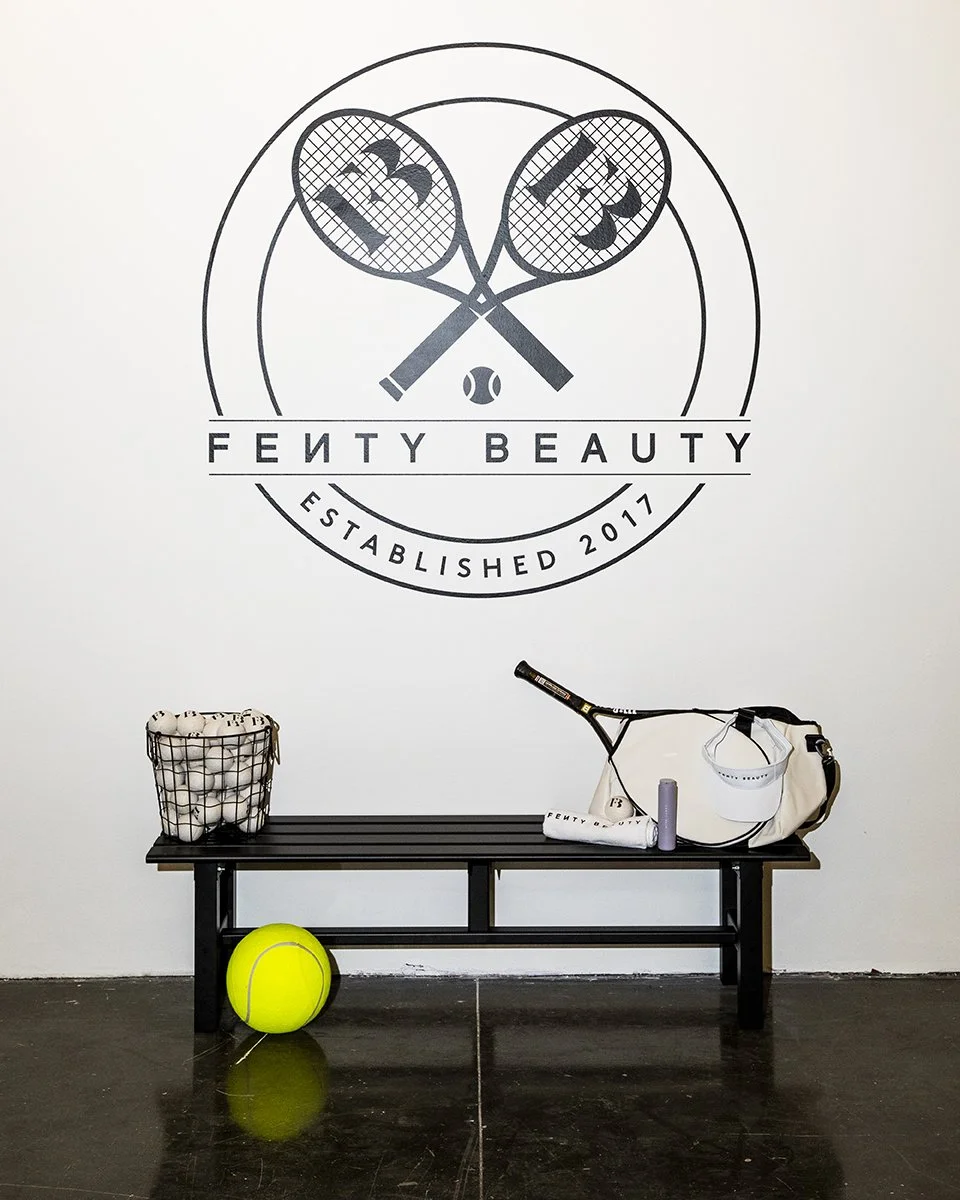 A black bench with tennis equipment, a large tennis ball on the floor, in front of a white wall with a Fenty Beauty logo and tennis-themed design, including tennis rackets and a ball.