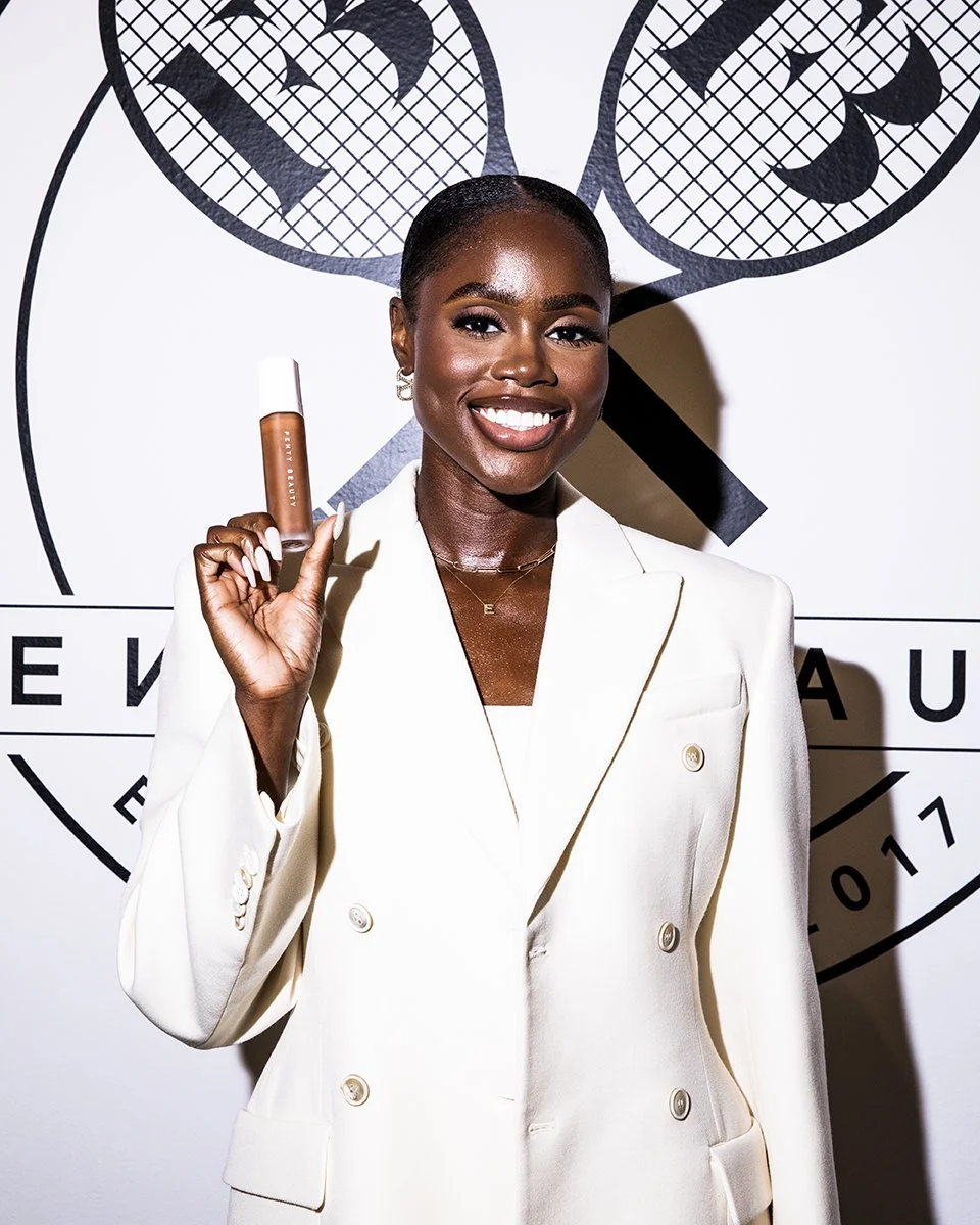 A smiling woman with dark skin and short hair holding a small bottle of makeup or skincare product. She is wearing a white blazer and standing in front of a designer logo background.