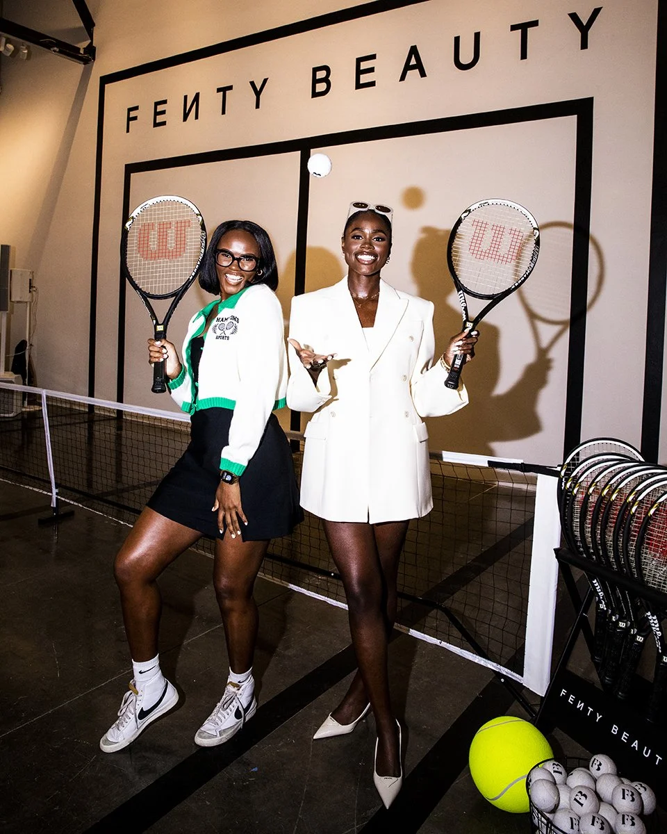 Two women with tennis rackets posing in front of a Fenty Beauty display at a tennis-themed event. One woman is wearing a white blazer dress with high heels, sunglasses, and holding a tennis racket. The other woman is dressed in a tennis-themed jacket