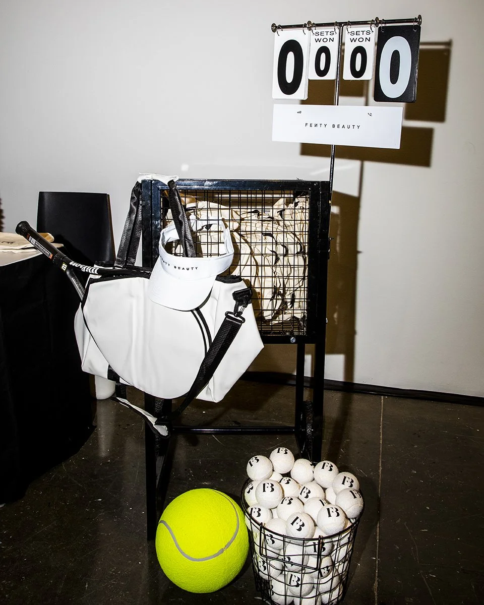 A ping pong scoreboard showing zeros, surrounded by tennis balls, a golf ball, and equipment, with a white Fenty Beauty bag hanging from a black metal stand, set against a plain white wall.