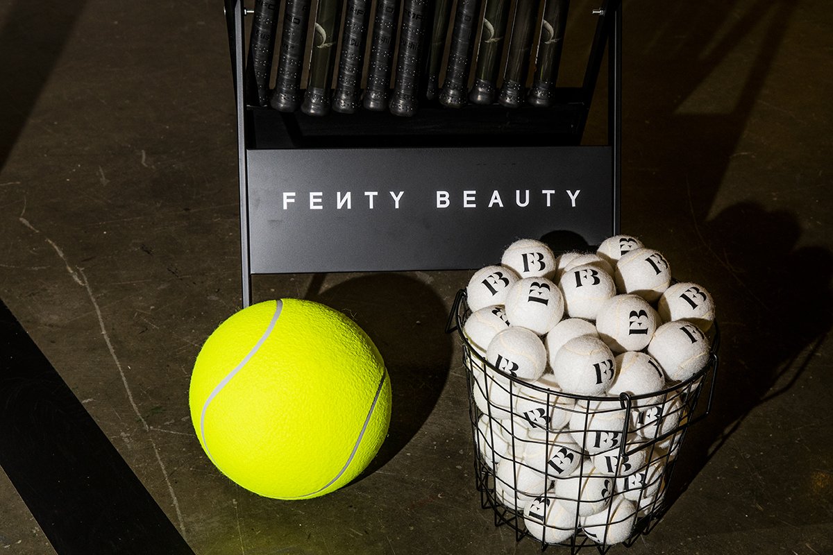 A large yellow tennis ball, a wire basket filled with white practice golf balls marked with 'FB', and a display stand with various tennis rackets behind it, all on a dark surface.