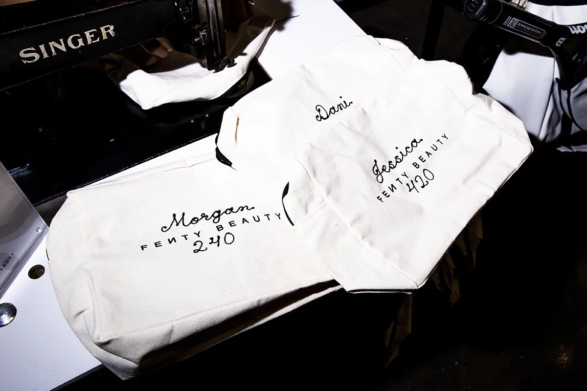 Two white tote bags with black cursive and text reading 'Morgan Fenty Beauty 240' and 'Dani Jessica Fenty Beauty 120' on a black surface, with a Singer sewing machine partially visible in the top left corner.