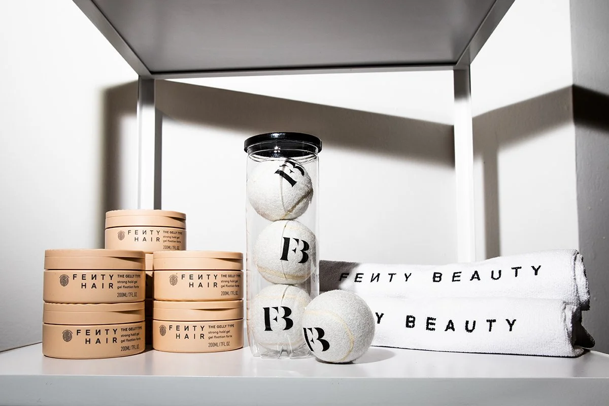 Fenty Hair styling products, a clear container with tennis balls marked with 'FB', and Fenty Beauty towels on a white shelf.