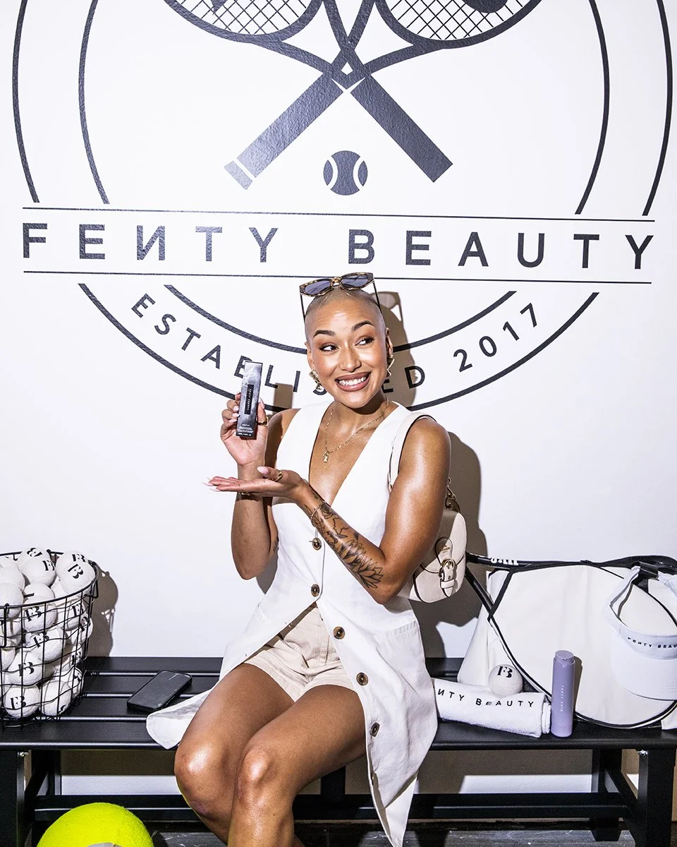 A woman with a shaved head and sunglasses on her head sitting in front of a Fenty Beauty logo on a wall, holding a beauty product, smiling, with a tennis ball and beauty products on a bench beside her.