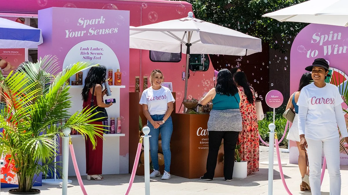 A group of women at a colorful outdoor event booth with pink and purple decor, promoting scented products. Some women are talking at the booth, while others are smiling and walking by. There are umbrellas, plants, and banners with phrases like 'Spark