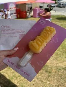 A person holding a pink flyer with a yellow, mustache-shaped dessert on a stick. There are tents and other people in the background outdoors.