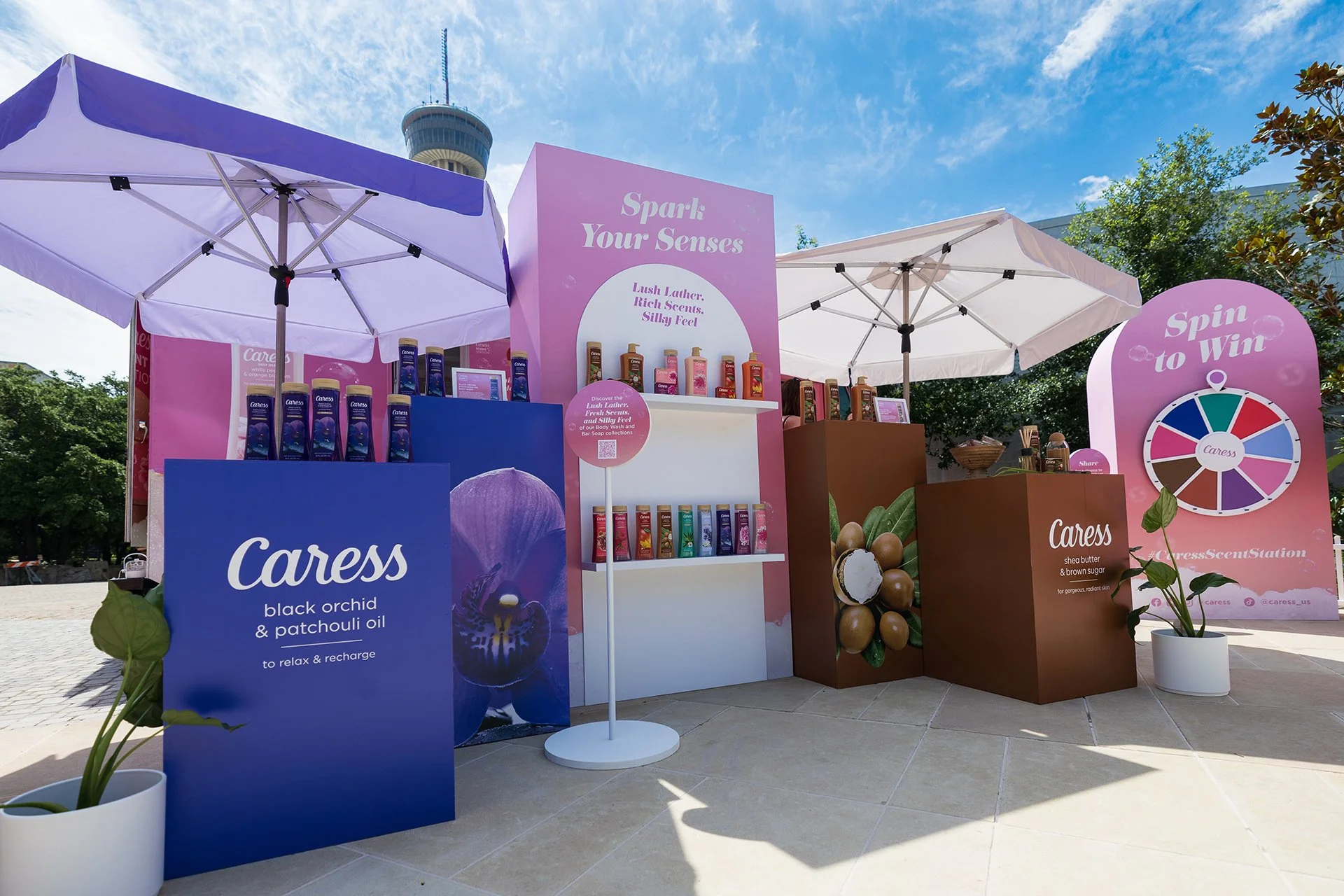 Outdoor display booth for Caress body wash and skincare products, featuring pink and brown color themes, with large informational signs, shampoo bottles, potted plants, and a 'Spin to Win' wheel, under blue sky with few clouds.