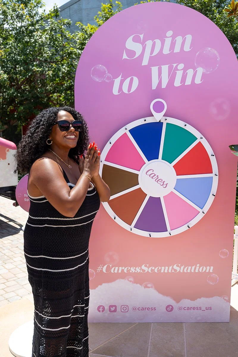 A woman in a black dress with white stripes standing outdoors next to a pink spinning wheel sign that says 'Spin to Win' and '#CaressScentStation'. She is smiling, wearing sunglasses and jewelry, with her hands clasped together, and has curly hair.