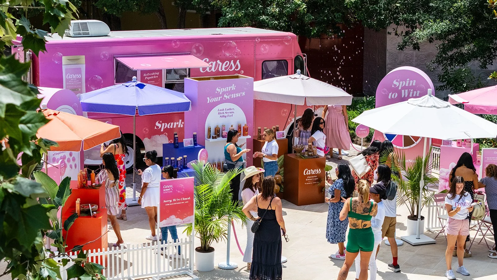 A busy outdoor event with a pink Caress-themed booth, pink and white umbrellas, and people browsing and engaging with products on display, surrounded by greenery.
