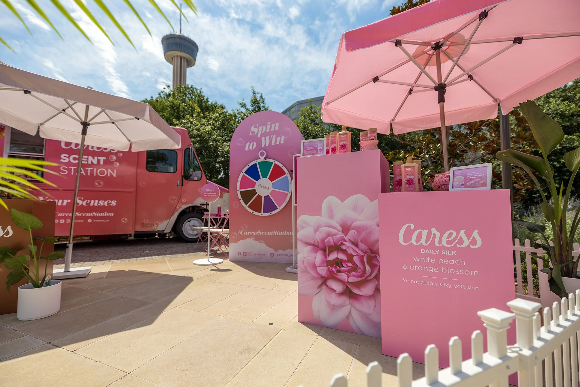 A pink mobile scent station truck with white text, two umbrellas providing shade, and a pink display booth for Caress daily silk body wash with a large flower image and product information, set outdoors under a partly cloudy sky.