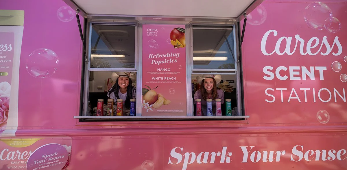 A pink mobile station titled 'Caress Scent Station' selling refreshing popsicles in flavors mango and white peach, with two smiling women in cowboy hats working inside the station.