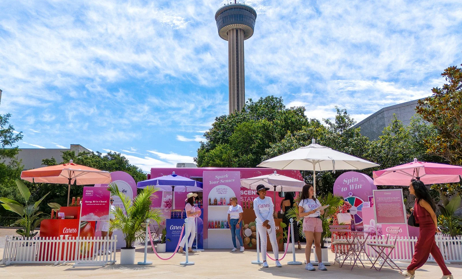 An outdoor event setup with pink and purple umbrellas, white picket fence, and a pink booth. People are standing and walking, some wearing white and pink clothing. A tall observation tower is visible in the background under a partly cloudy sky.