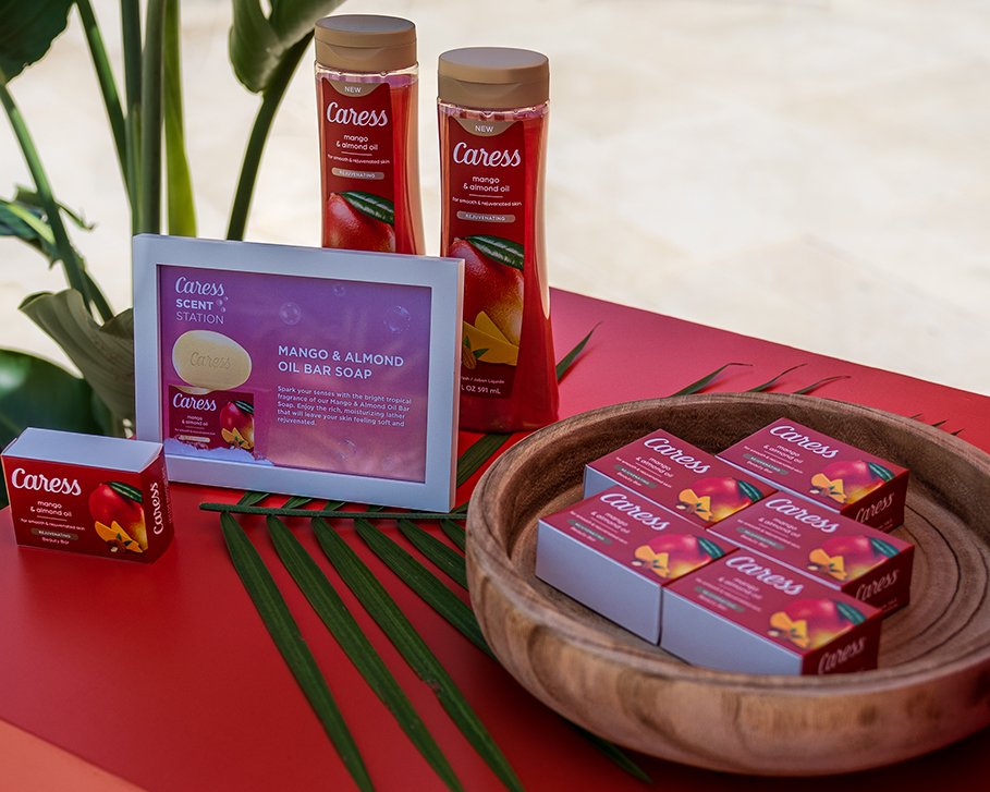 Display of Caress Mango & Almond Oil products, including soap bars and bottles, arranged on a table with a wooden bowl and green leaves for decoration.