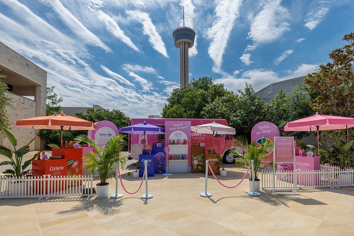 Outdoor event setup with colorful booths, umbrellas, potted plants, and a pink truck, with the San Antonio Tower in the background under a partly cloudy sky.