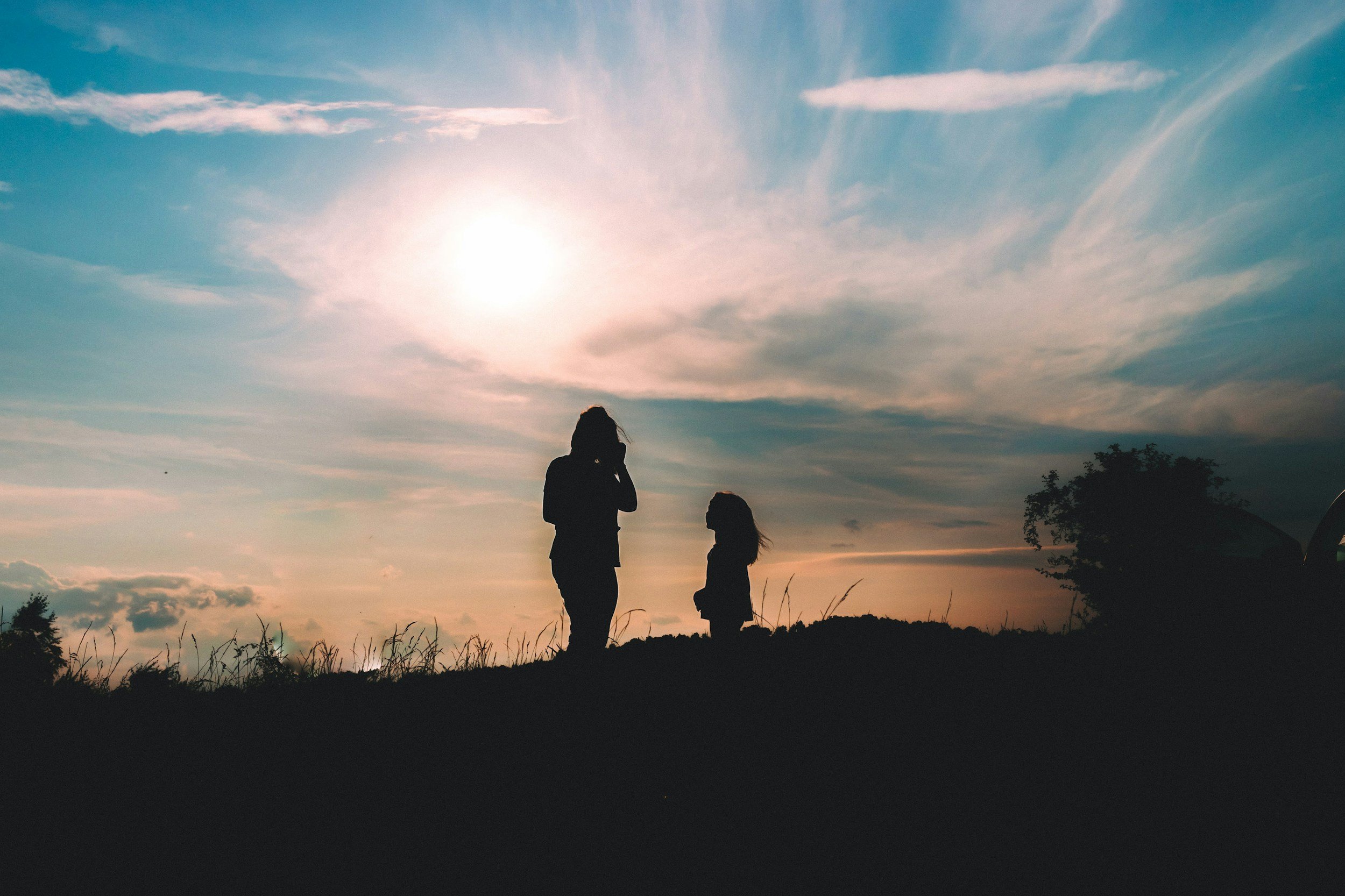Silhouettes of two women standing on a grassy hilltop, watching the sunset with clouds in the sky.