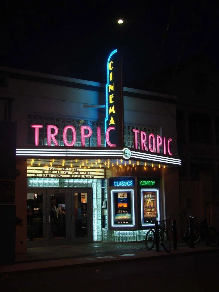Night view of the Tropic Cinema with neon signs displaying 'Tropic' in pink and 'Cinema' in yellow, and posters indicating 'Classics' and 'Comedy' screenings.