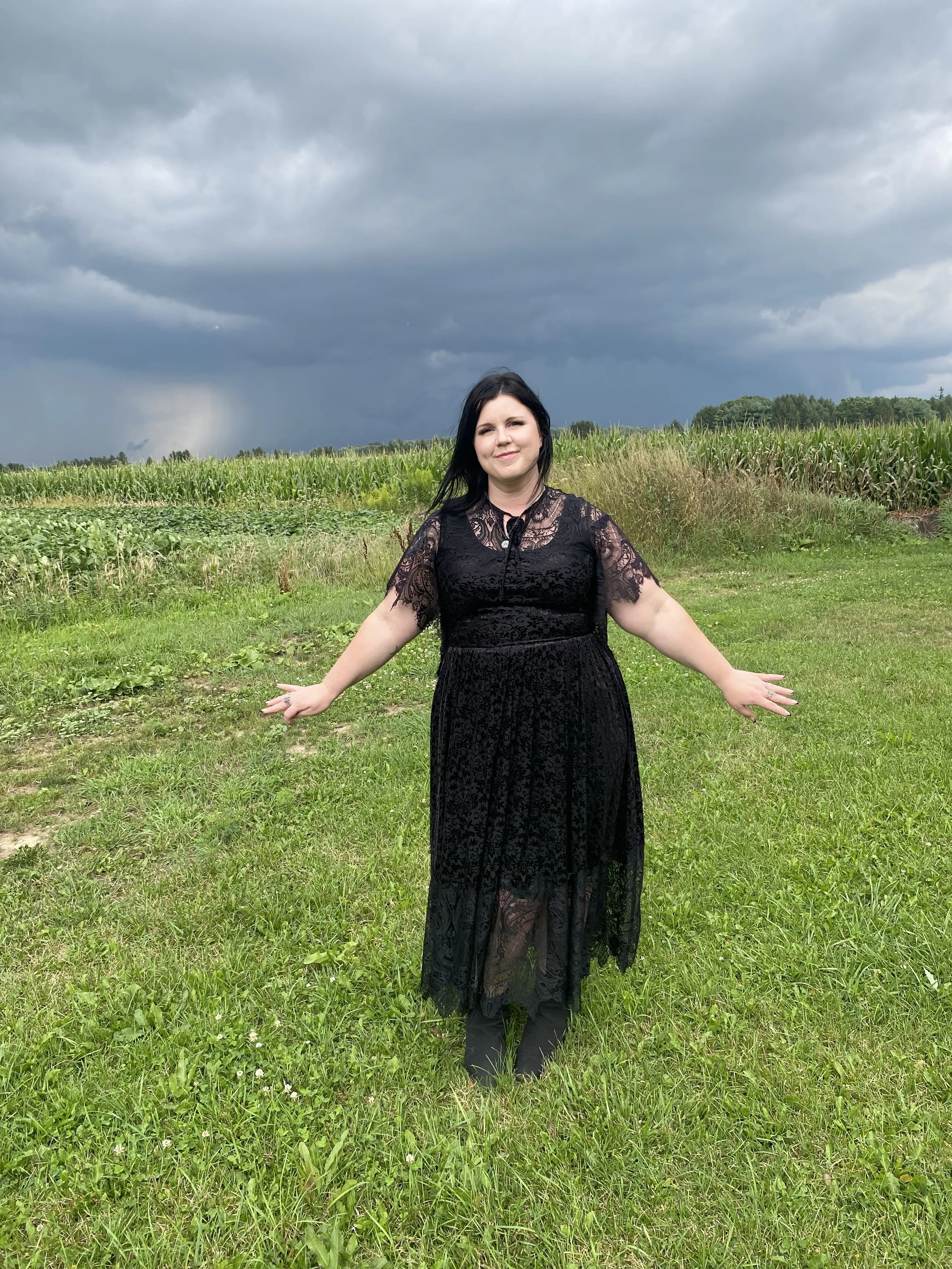 A woman standing in a grassy field with her arms outstretched, wearing a black lace dress, with a cloudy stormy sky and cornfields in the background.