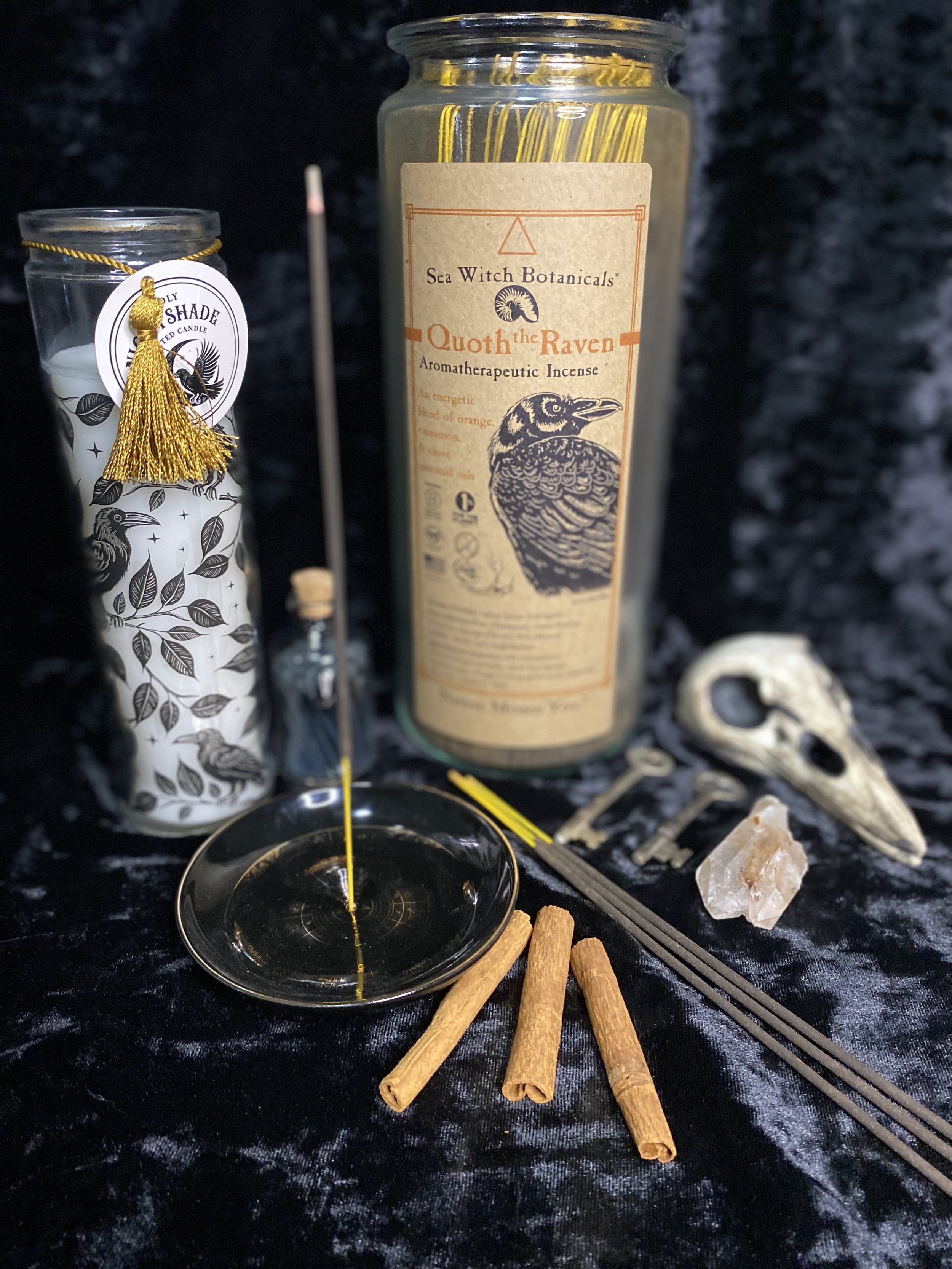 A collection of spiritual and ritual items including a tall large candle labeled 'Sea Witch Botanicals Quoth the Raven,' a white candle with a black bird illustration and gold tassel, a small vial, a black and gold mirror dish with a yellow candle wick, three cinnamon sticks, a black and gold and gray card reading 'tarot,' a clear quartz crystal, and a bone or shell object, all arranged on black fabric with a dark background.