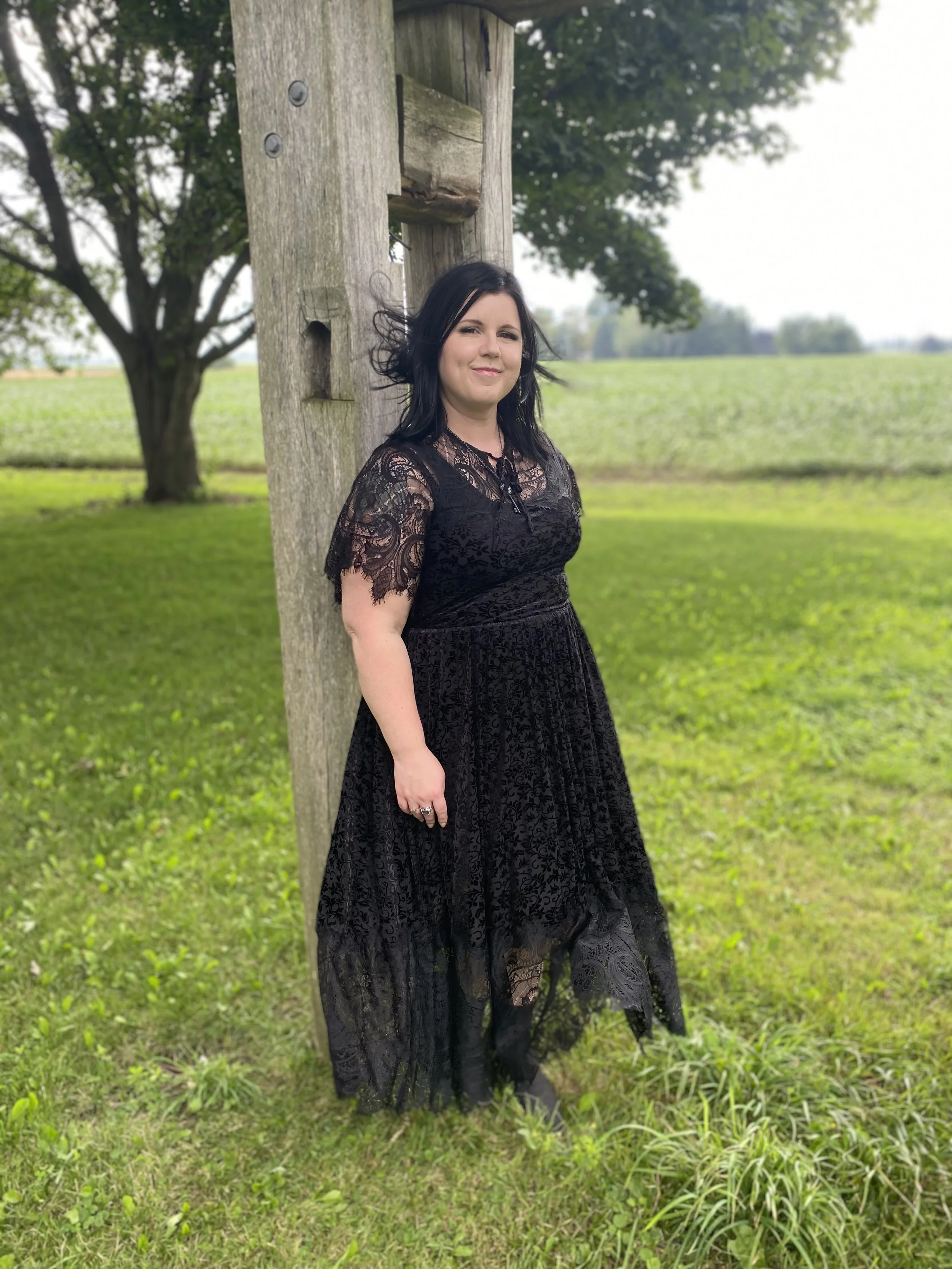 A woman with dark hair and a confident expression standing next to a weathered wooden utility pole outdoors in a lush green field with trees in the background, wearing a black lace dress.