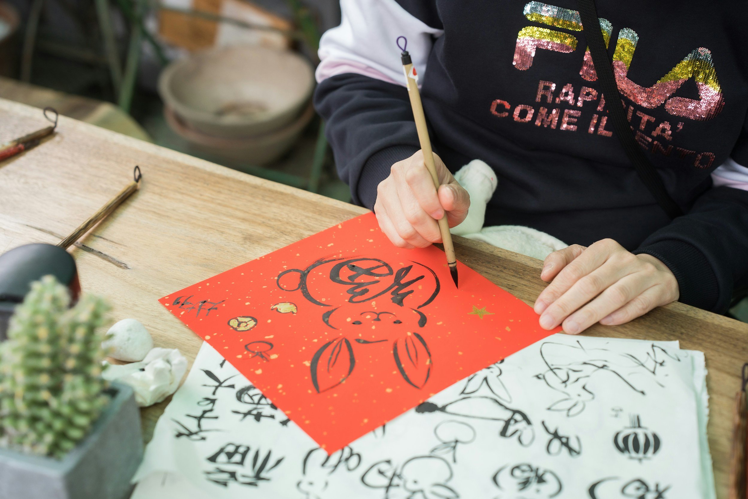 Person writing with a brush on festive red paper with a pumpkin illustration. Various sketches and calligraphy papers are scattered on a wooden table, with some plants visible in the foreground.
