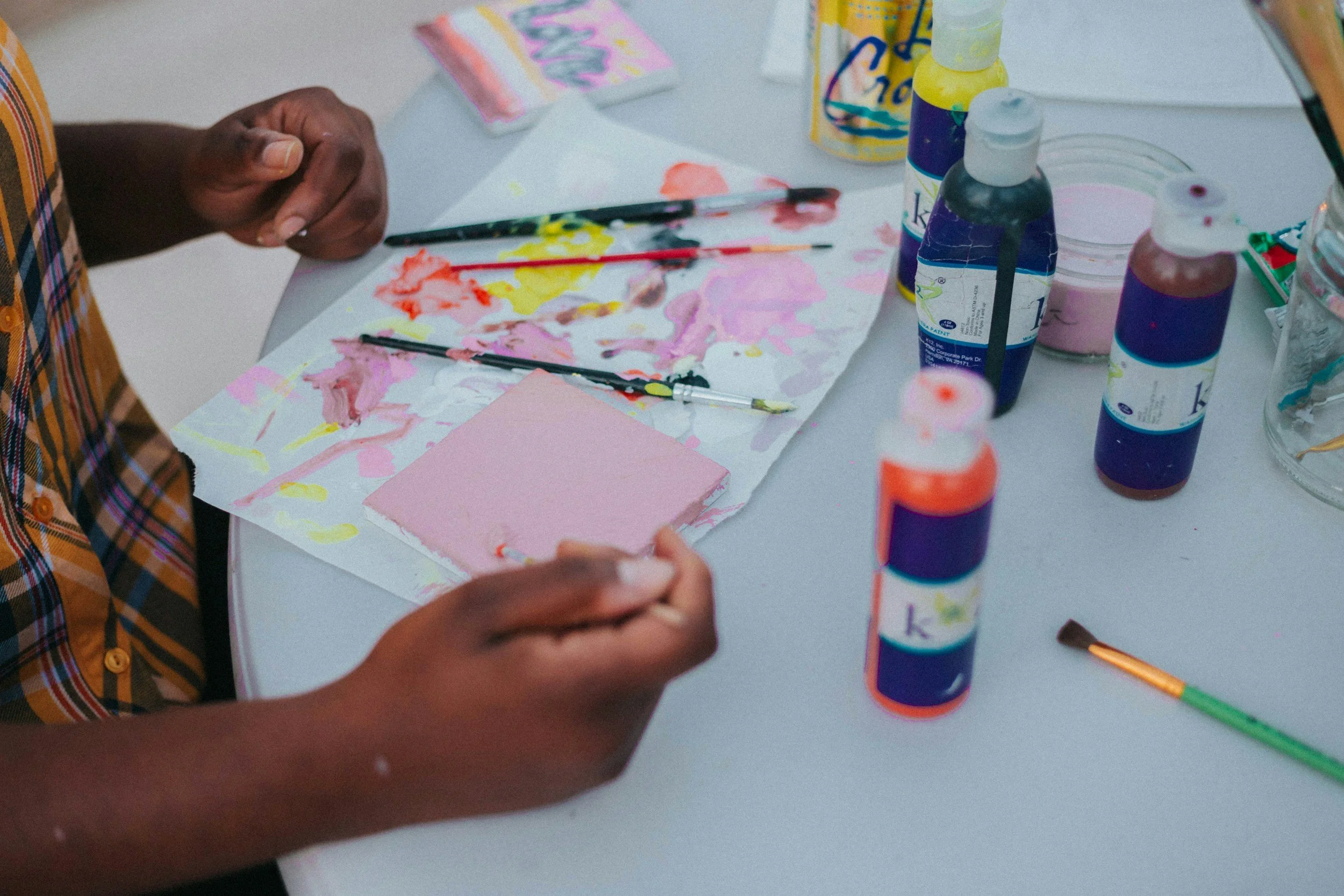 Close-up of a child's hands working on a craft project with watercolor paints, brushes, and a pink sponge, surrounded by colorful bottles of paint on a white table.