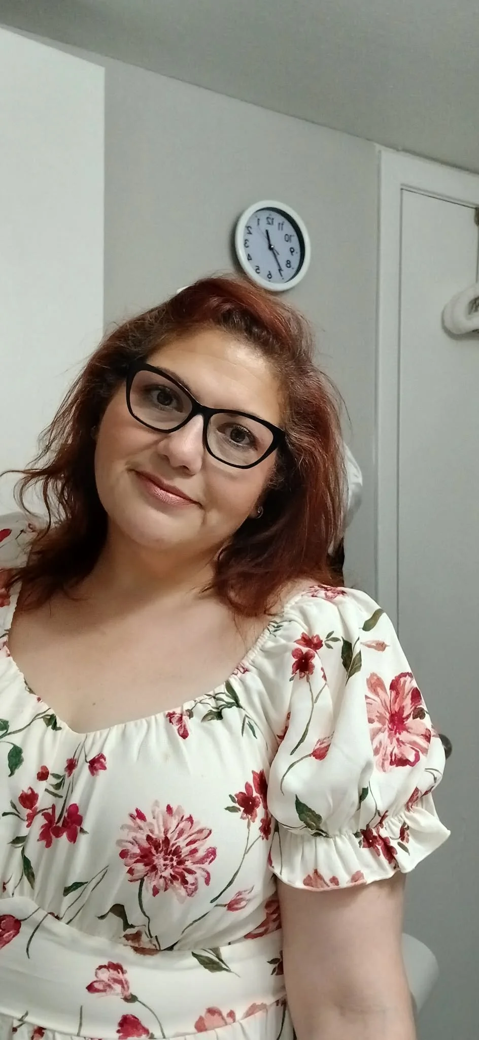 Smiling, female teacher wearing a floral dress and glasses with red hair and her head tilted to the right side.
