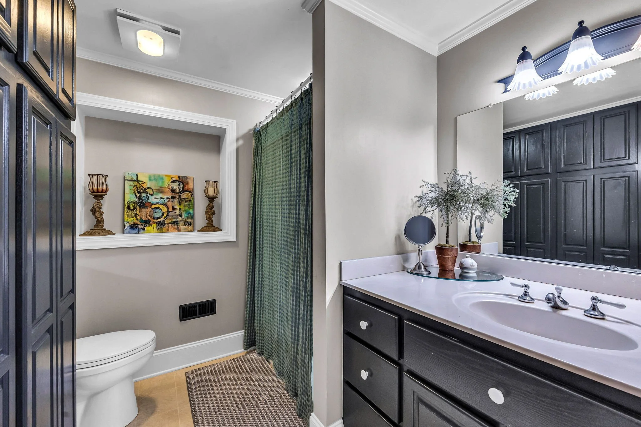 A bathroom with a white toilet, a dark blue cabinet, a white vanity with a mirror, decorative plants, a colorful abstract painting, a green shower curtain, and ceiling lights.