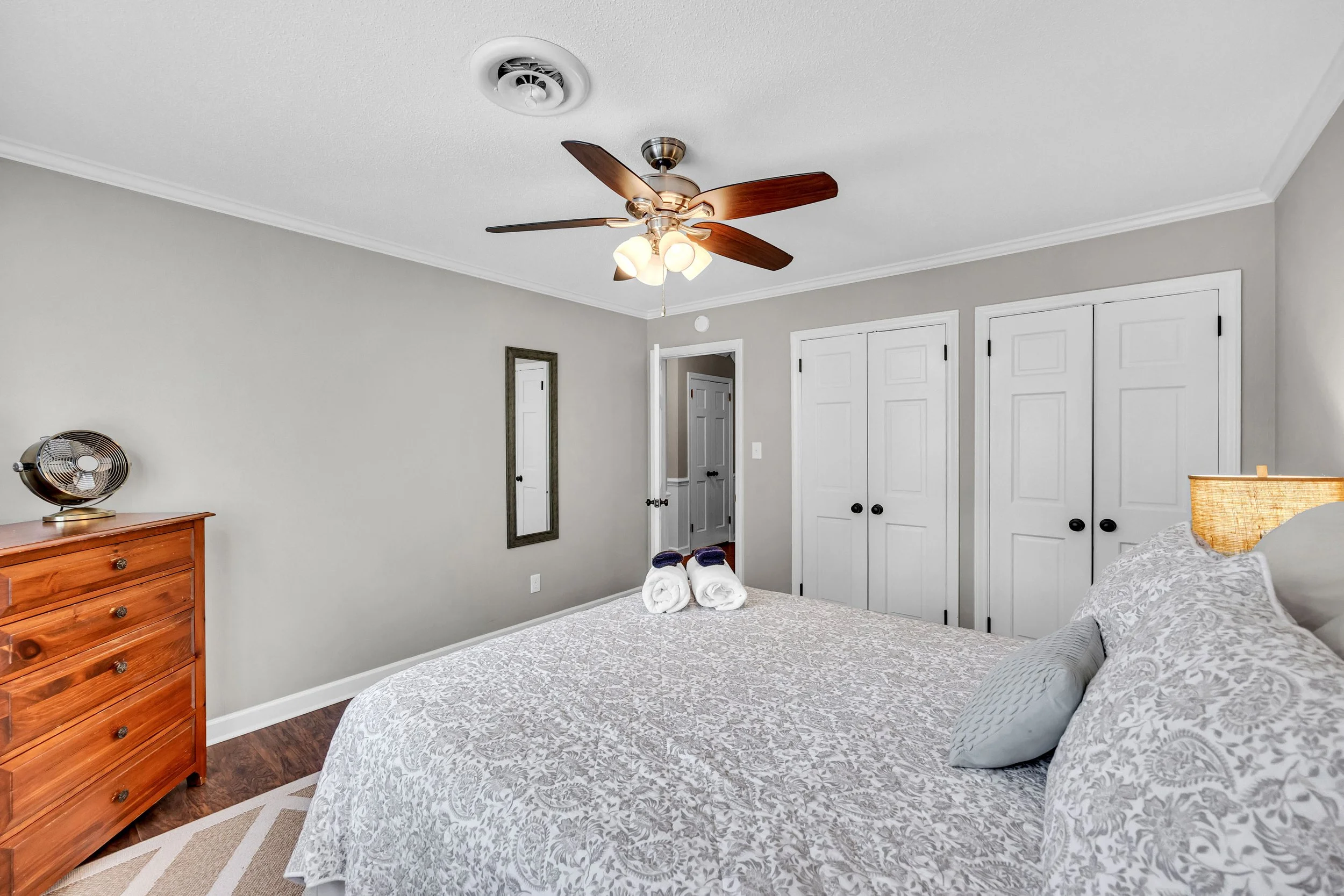 A bedroom with a bed covered in a light patterned comforter, pillows, and towels on top. There is a wooden dresser with a fan on it, a mirror on the wall, and white closet doors. The room has gray walls, a ceiling fan with lights, and hardwood floors.