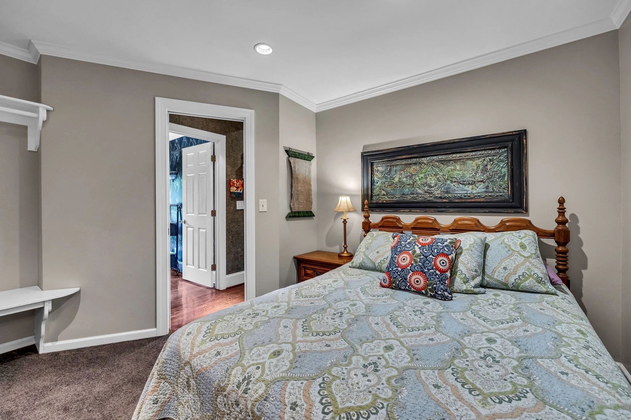 A cozy bedroom with a wooden bed and colorful pillows, a bedside lamp, gray walls, a framed artwork above the bed, and a doorway leading to another room.