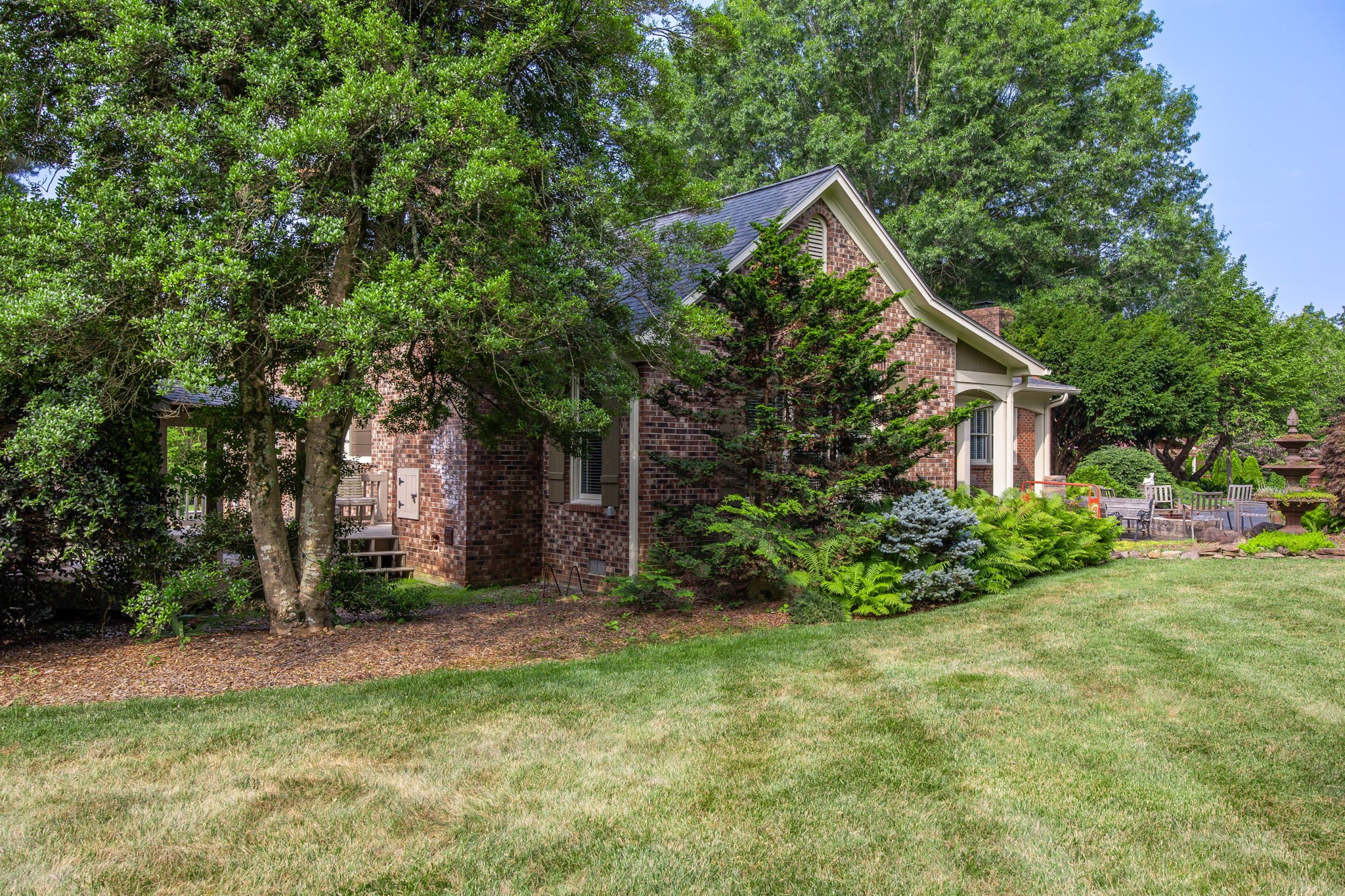 A backyard with green grass, trees, and shrubs surrounding a brick house with a grey roof.