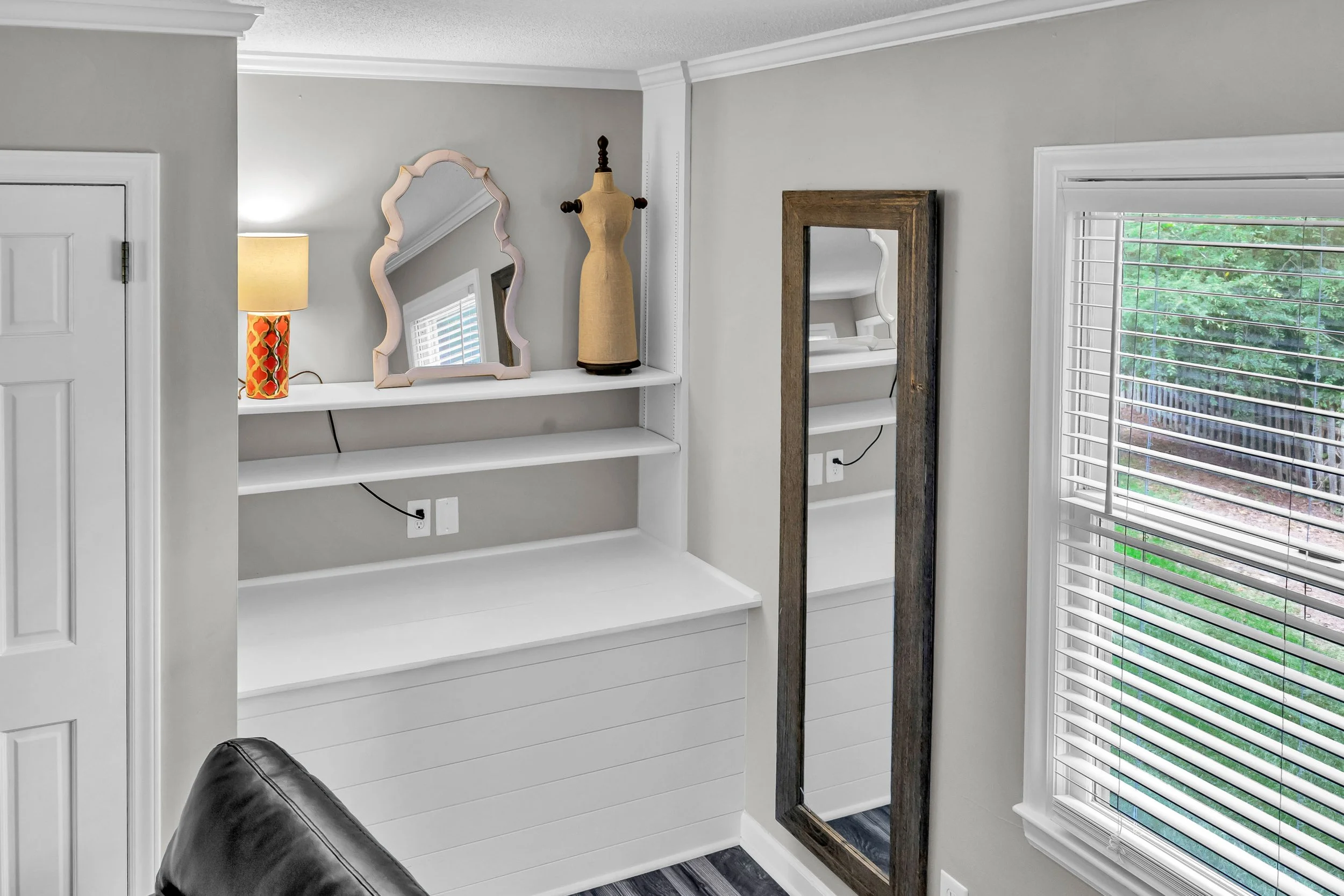 Interior of a bright room with white walls, a large window with white blinds, a tall wooden mirror, a mannequin, and a white shelf with decorative items, a lamp, and electrical outlets.