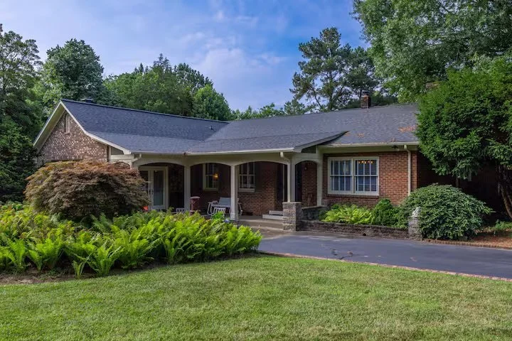 Front view of a brick house with a large front porch, surrounded by lush green landscaping and tall trees, on a clear day.